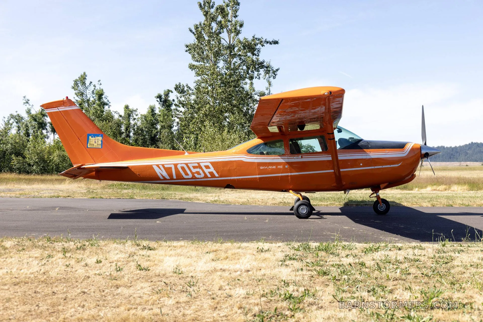 A bright orange Cessna 182 Skylane 50 with white stripes and black accents is parked on an asphalt strip next to a grassy field with trees in the background.