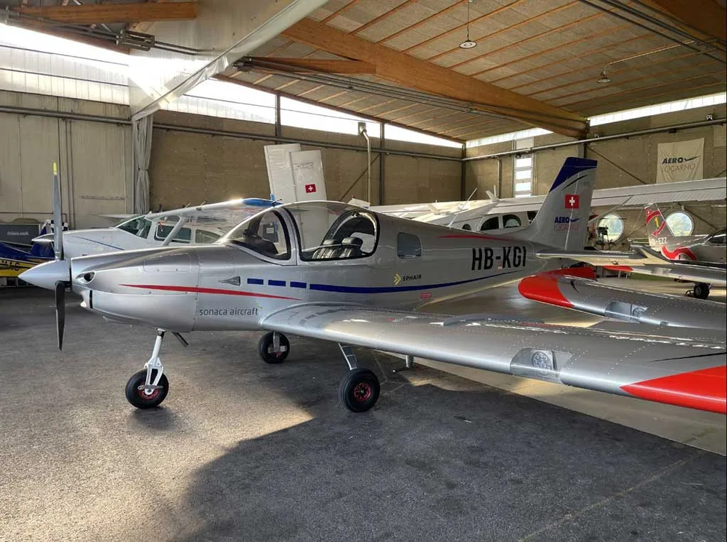 A silver Sonaca 200 aircraft with blue and red stripes, registration HB-KGI, parked inside a hangar, viewed from the front-right.