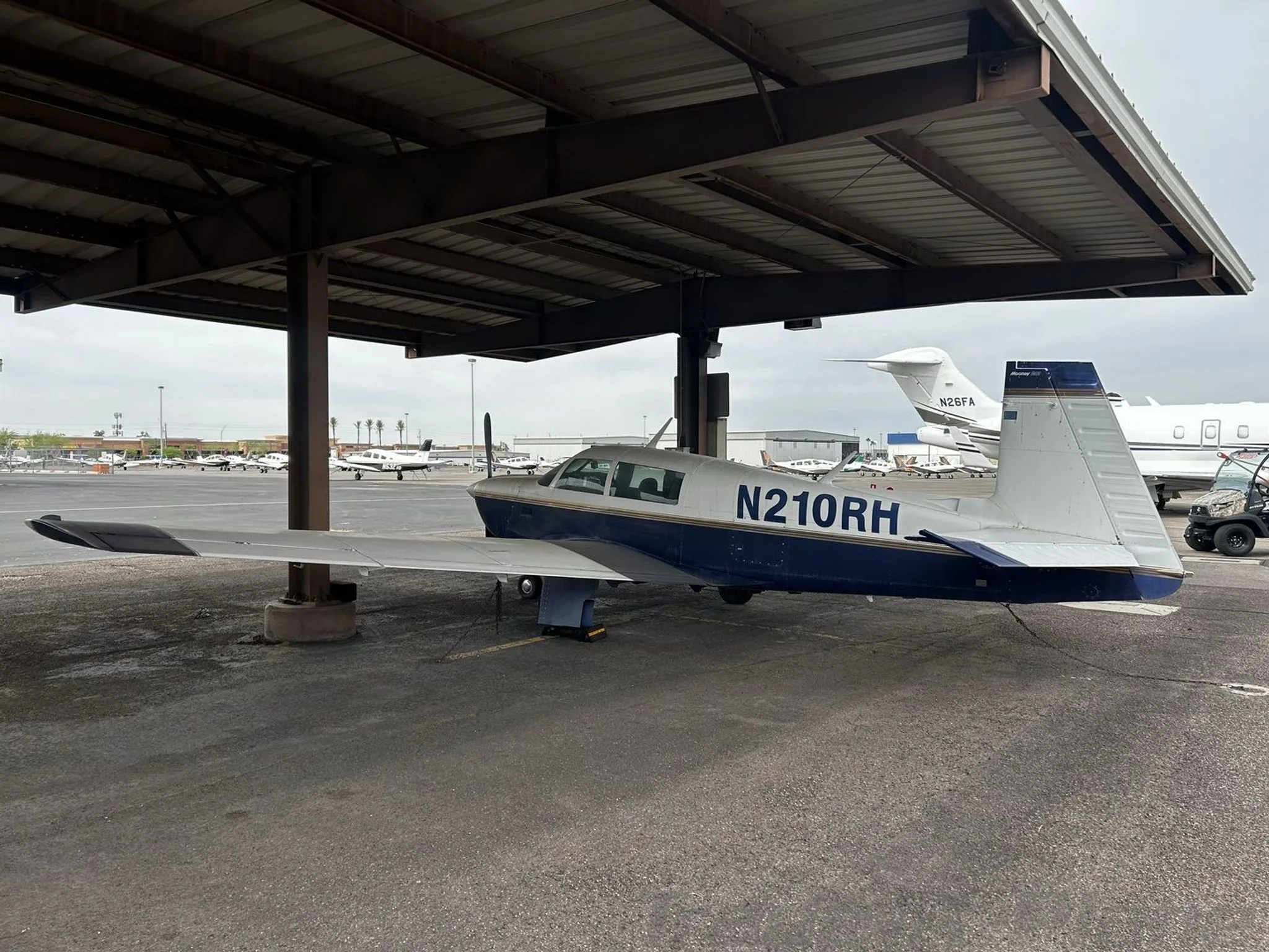 Side view of the blue and white single-engine aircraft N210RH parked under a covered shelter with other aircraft in the background.