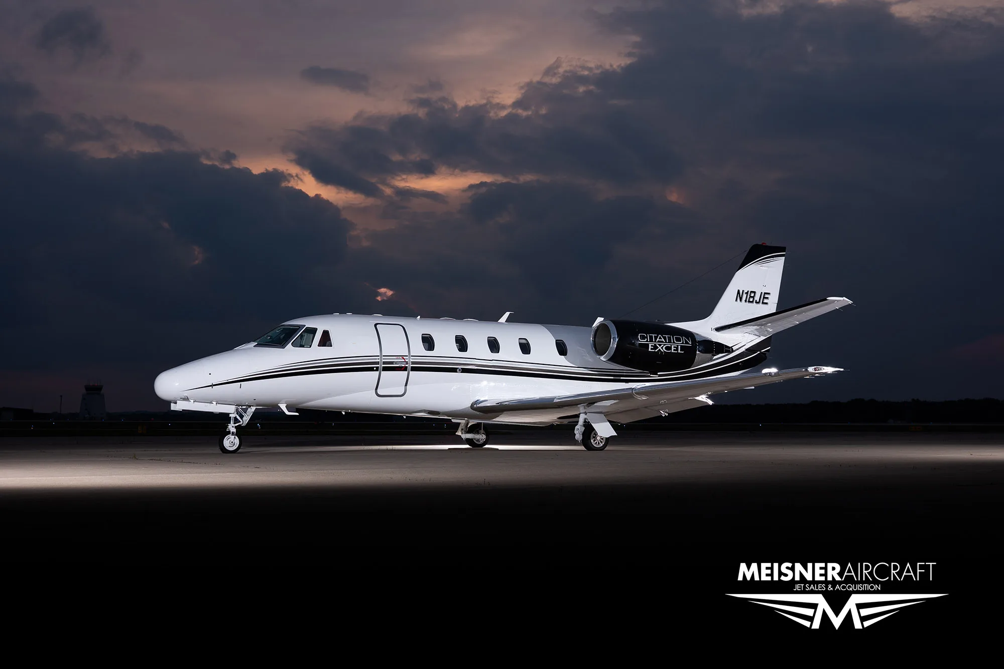A white Citation Excel business jet with black and white stripes on the tail and engines, parked on a tarmac at dusk.