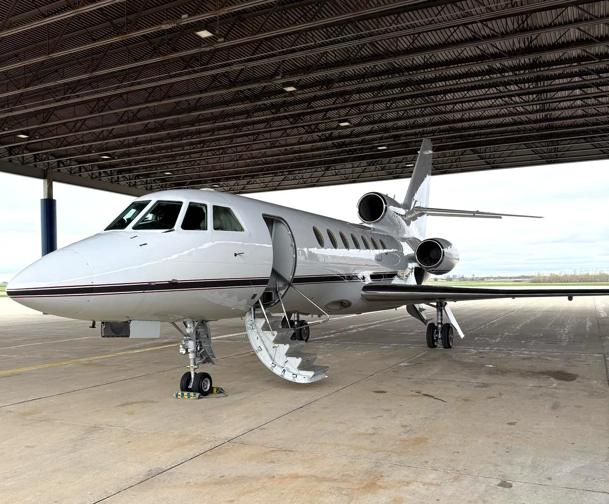 A front-left view of the white Dassault Falcon business jet with its airstairs deployed, parked under a hangar.
