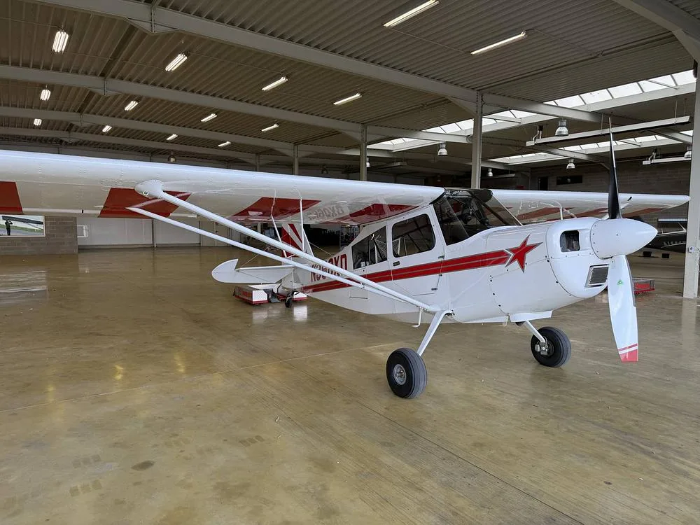 A white single-engine aircraft with red stripes and a red star on the nose, parked inside a hangar.