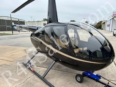 A black Robinson R44 Raven II helicopter with gold pinstripes is parked on a tarmac, viewed from the front-right.