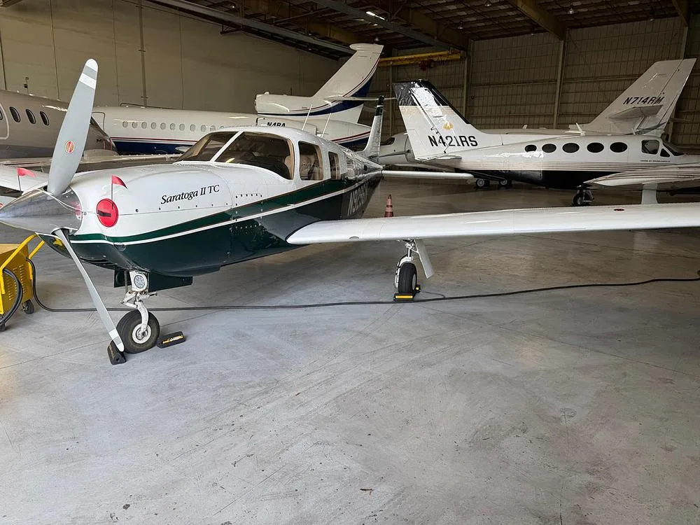 A Piper Saratoga II TC in dark green and white, parked inside a hangar with other aircraft.