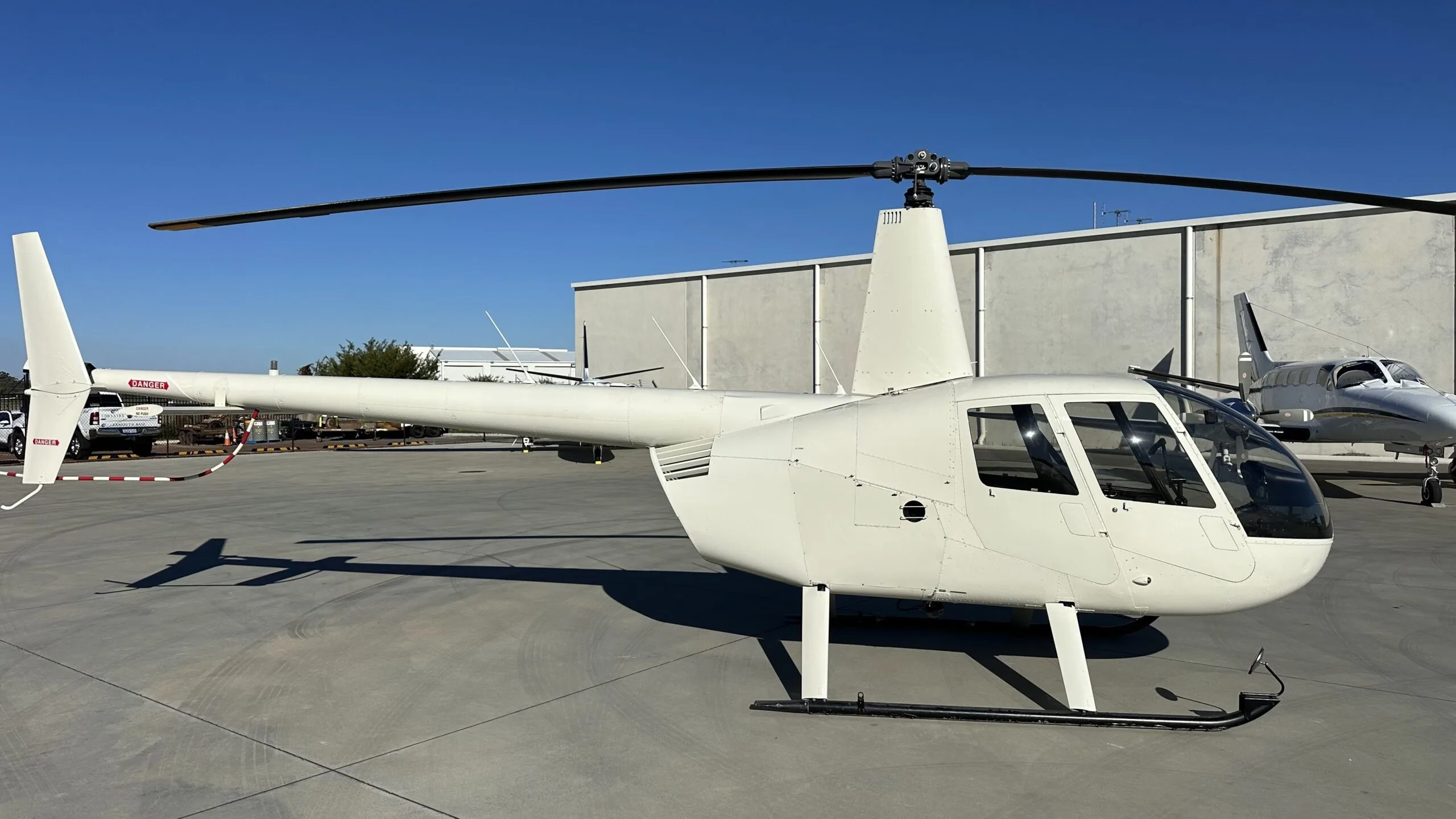 A white Robinson R44 helicopter parked on an airfield tarmac with hangars in the background.