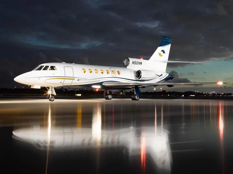 A white private jet with blue and gold accents is parked on a wet tarmac at night, reflecting its lights.