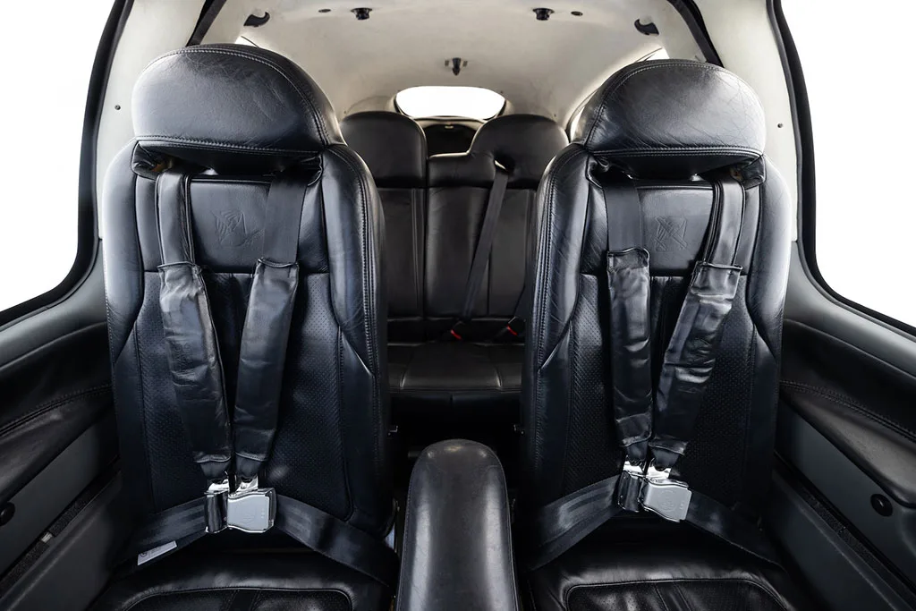 View from the rear of the aircraft cabin showing two black leather front seats with harnesses and two rear seats.
