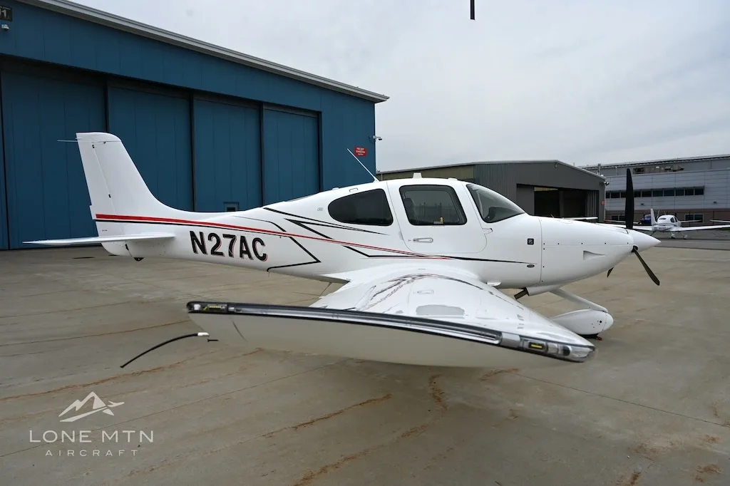 White Cirrus SR22 aircraft with red and black stripes, tail number N27AC, parked on an apron.