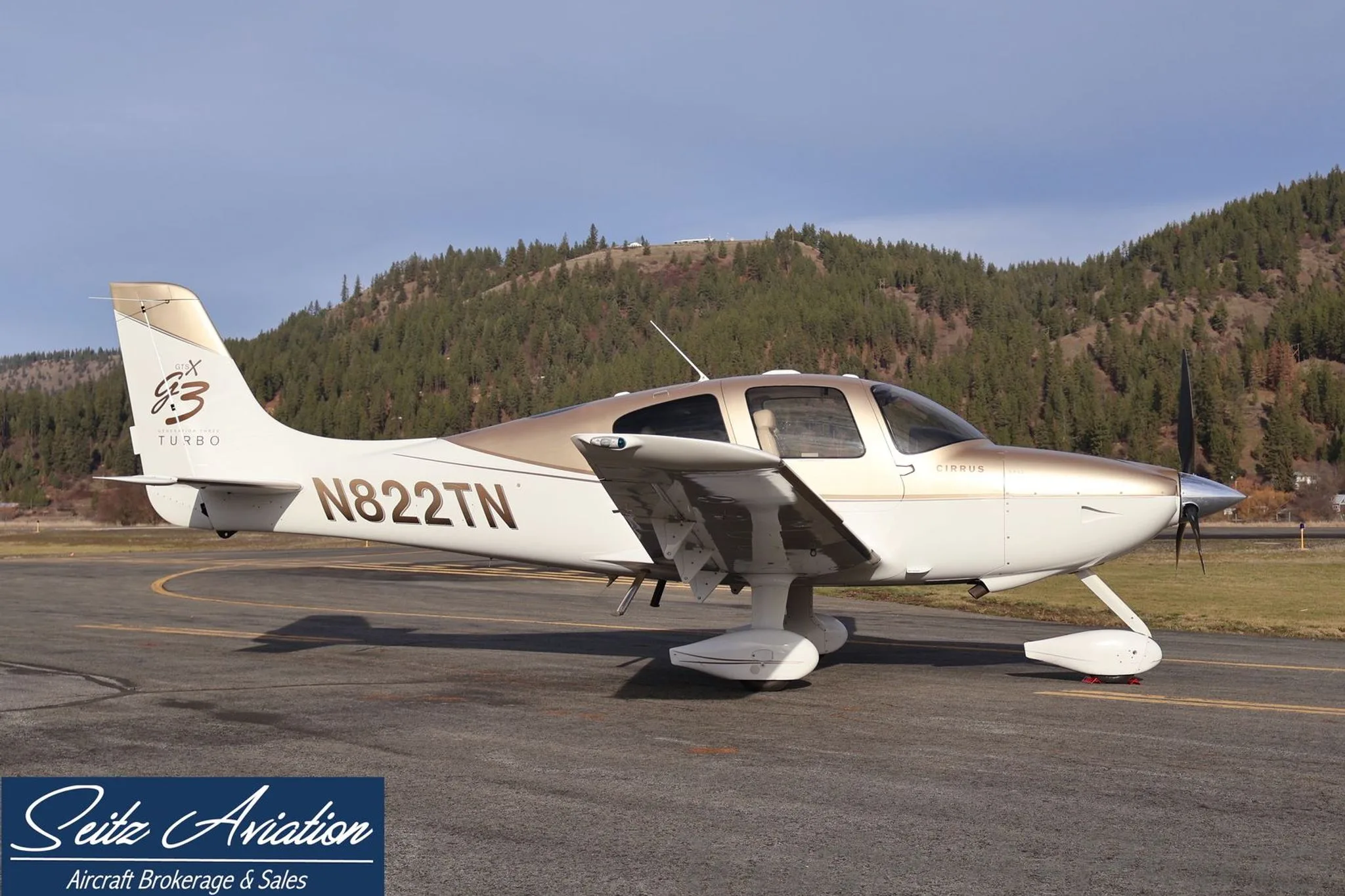 Side view of the Cirrus SR22T aircraft N822TN, painted white and gold, parked on an asphalt ramp with a forested hill in the background.