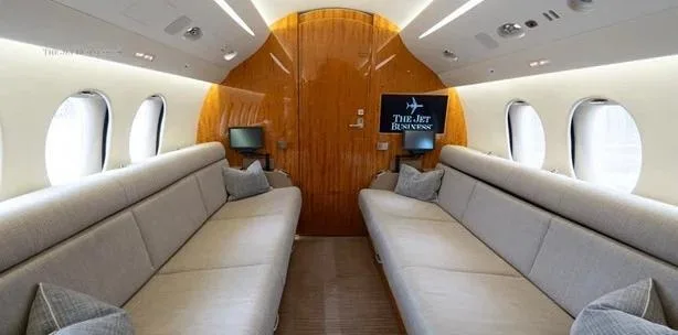 A private jet cabin interior featuring two long beige couches facing each other, with a wooden bulkhead at the end.