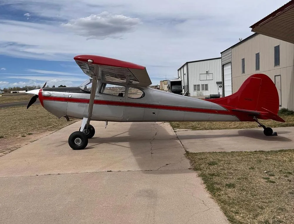 A silver and red Cessna 170B aircraft with a tailwheel, viewed from the side on an airfield tarmac.