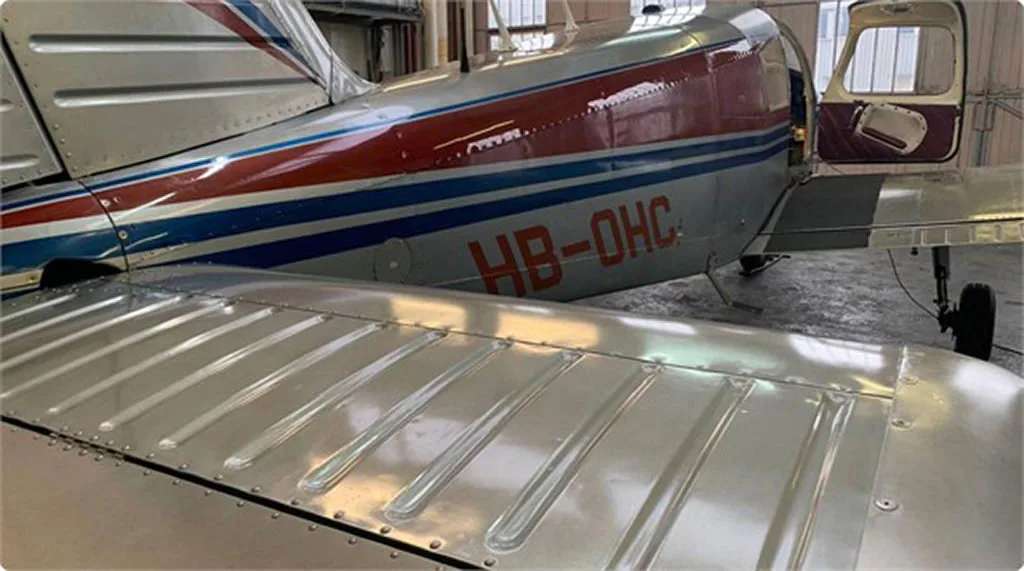 View of the wing and rear fuselage of a red and silver aircraft with blue stripes and registration HB-OHC, with an open door.