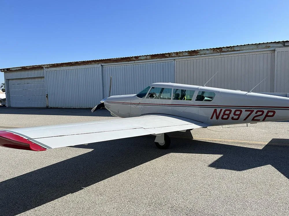 A side view of the white Piper Cherokee 180 (N8972P) with red and grey stripes parked on an asphalt ramp next to a hangar.