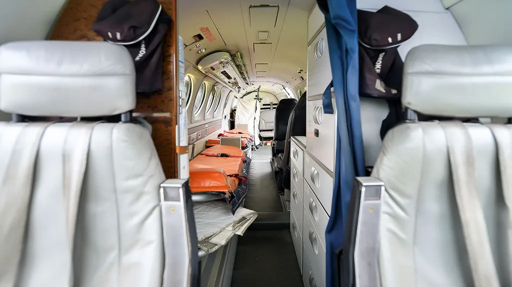 A view down the narrow aisle of an air ambulance cabin, showing multiple orange stretchers and white seats.