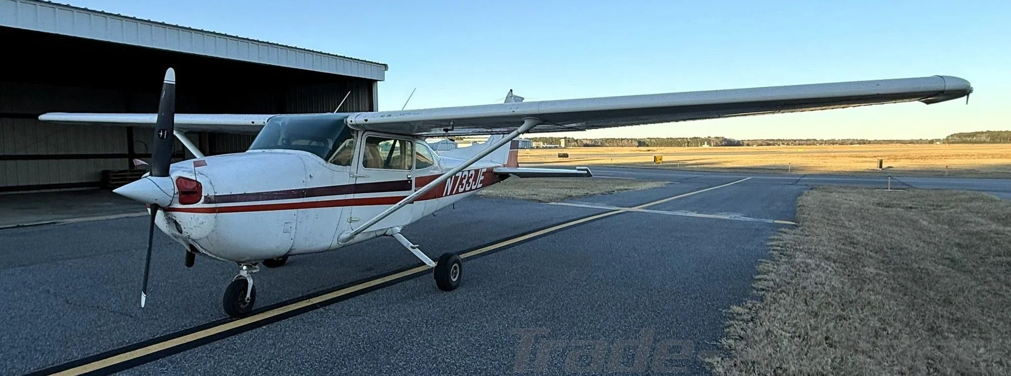 A white Cessna 172N with red and black stripes, registration N733JE, parked on an airfield next to a hangar.