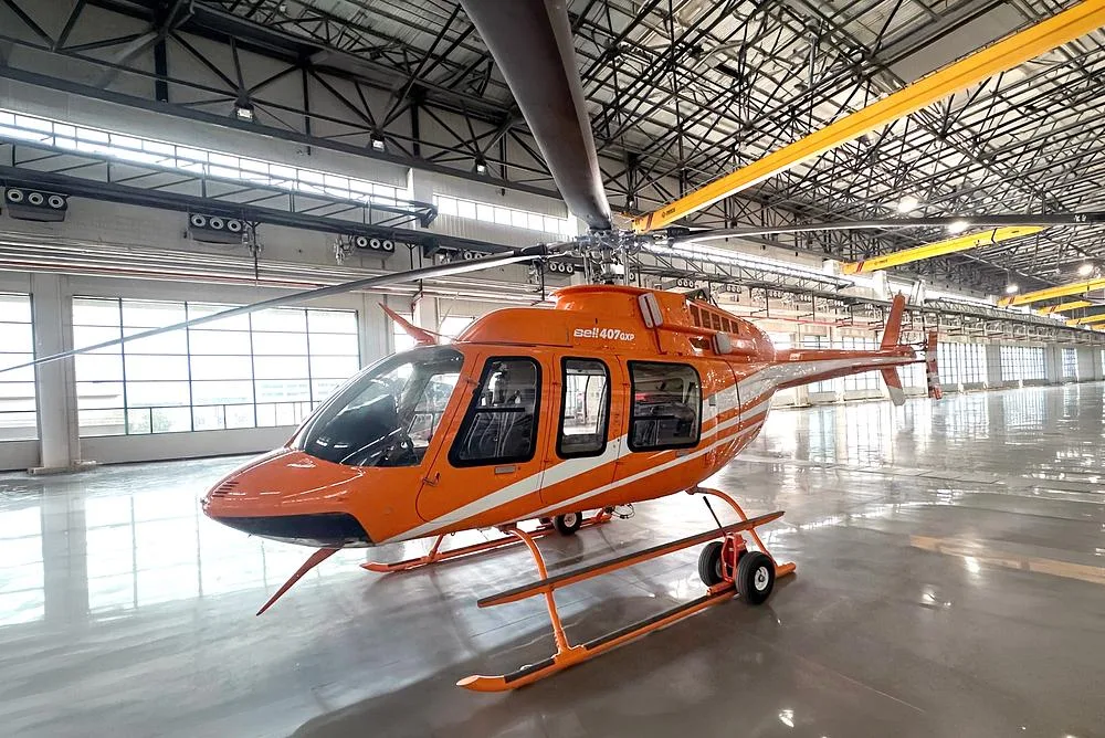 A Bell 407GXP helicopter in orange and white livery, parked inside a large hangar.