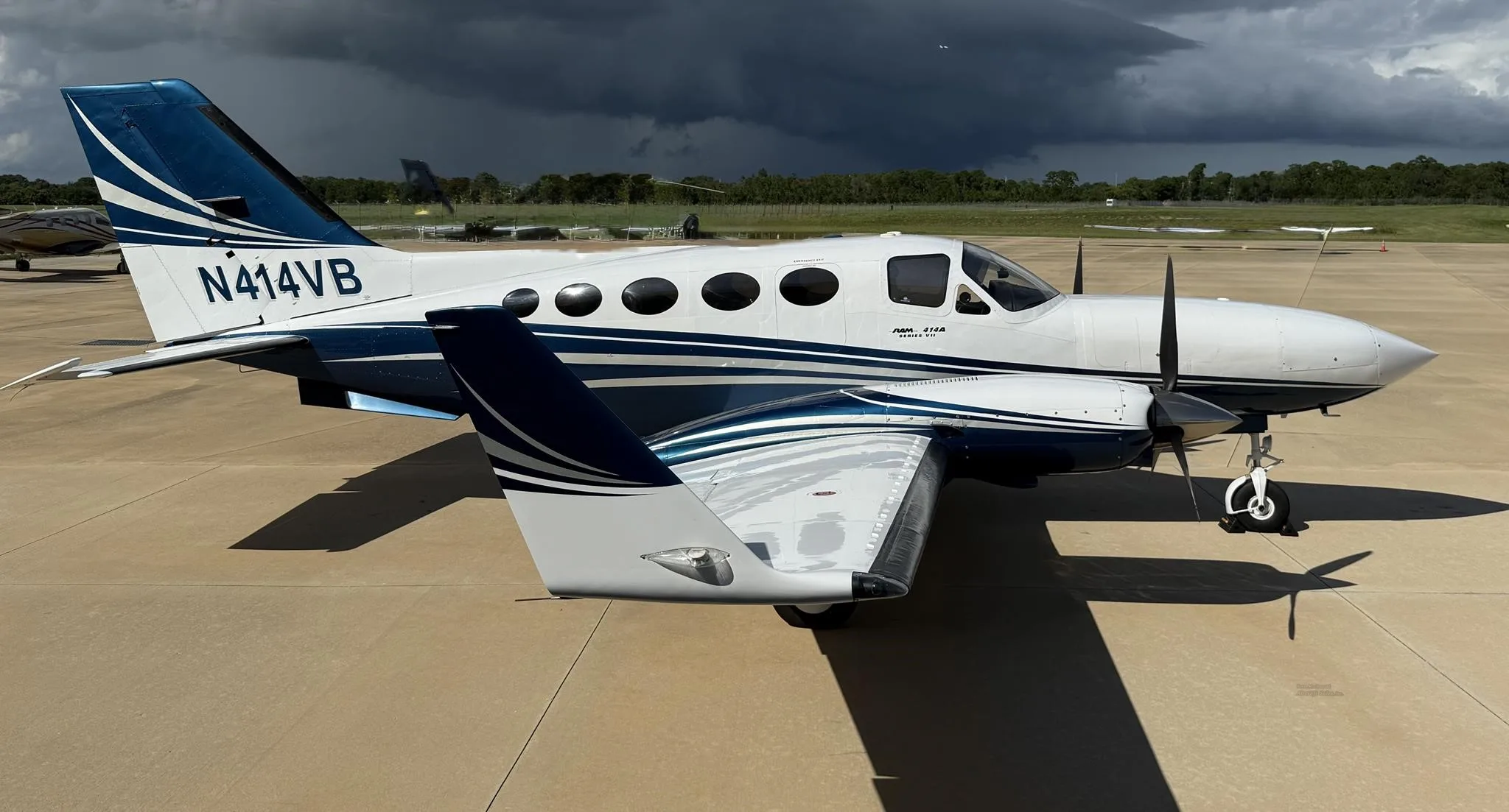 A white and blue Cessna 414 Chancellor twin-engine aircraft with tail number N414VB parked on an airfield under a cloudy sky.