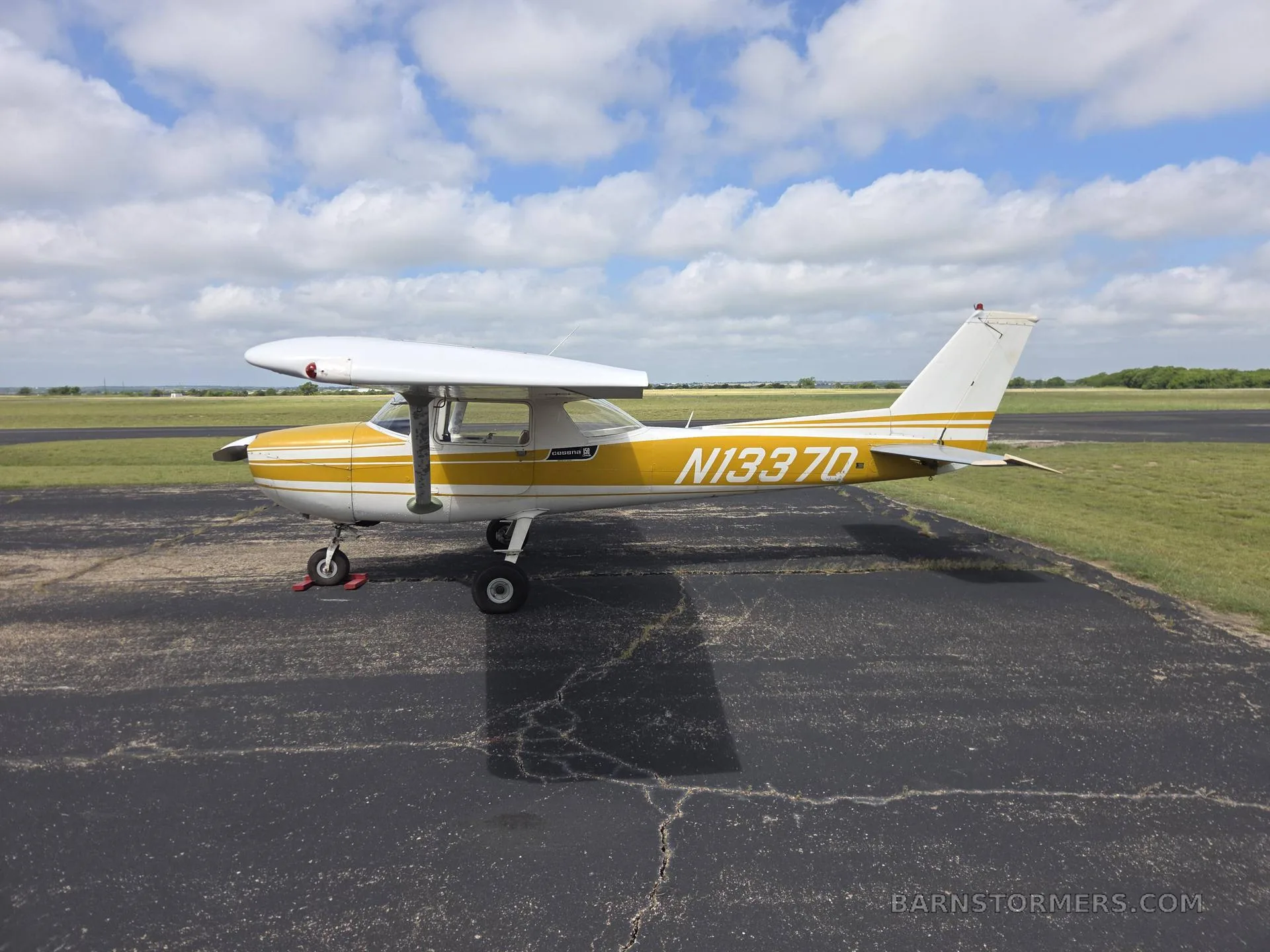 A white and yellow light aircraft (N13370) with a high wing, parked on an asphalt ramp, viewed from the left side.