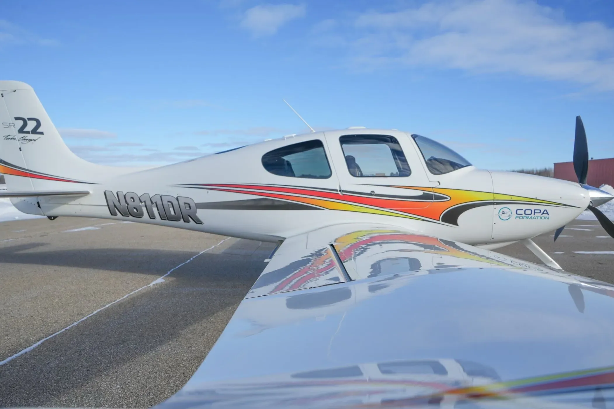 A white Cirrus SR22 aircraft with orange and red stripes parked on a snowy airfield, viewed from the wing.