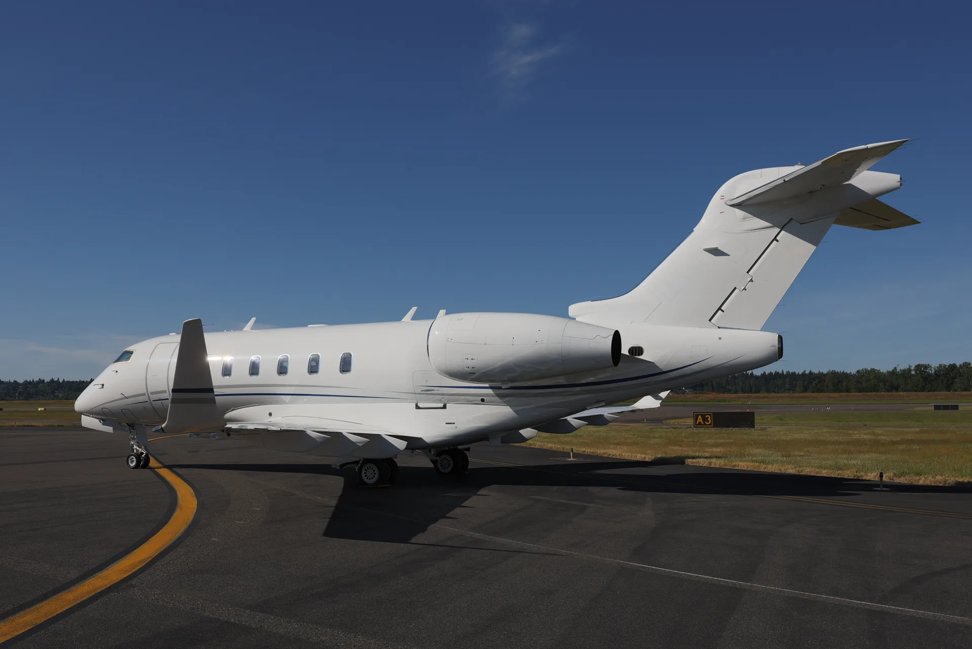 A white Bombardier Challenger business jet parked on an airport tarmac under a clear blue sky.
