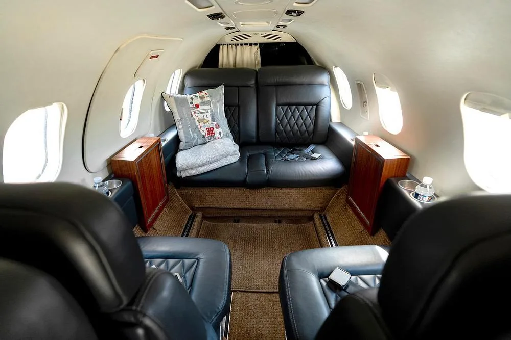 View of the aircraft cabin with two forward-facing black leather seats and a rear couch with a decorative pillow.