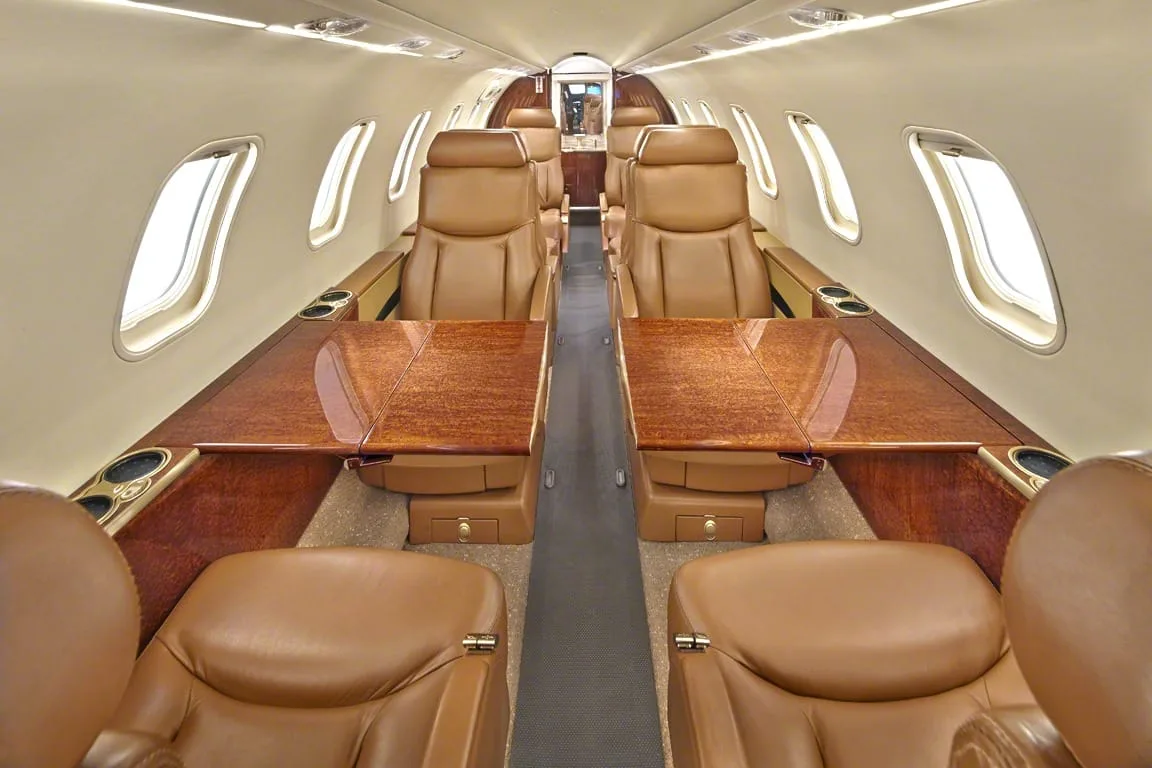 The interior cabin of a private jet with tan leather seats and polished wood tables.