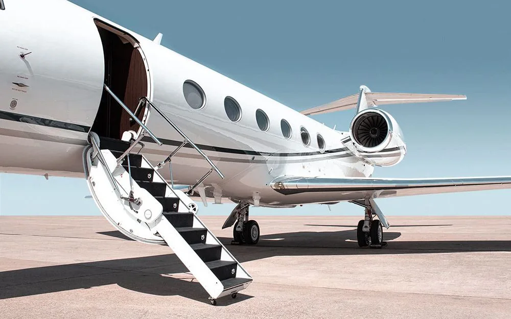A white private jet with its main door open and airstairs extended, parked on an airfield under a clear sky.