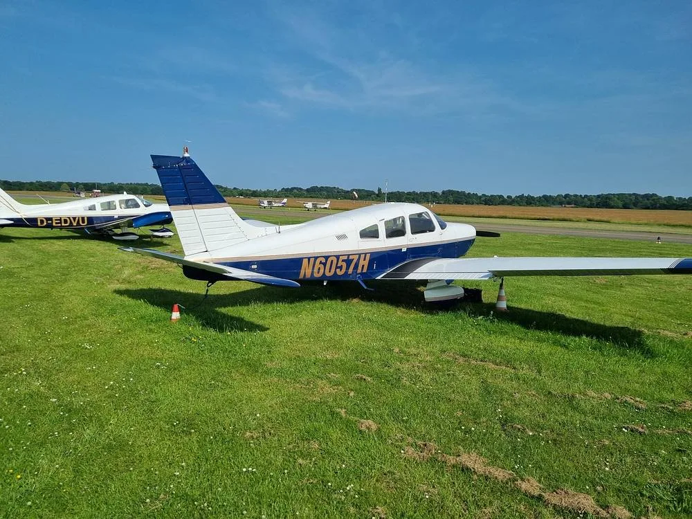 A Piper PA-28 Cherokee aircraft, registration N6057H, with blue and white paint, parked on grass next to another aircraft at an airfield.