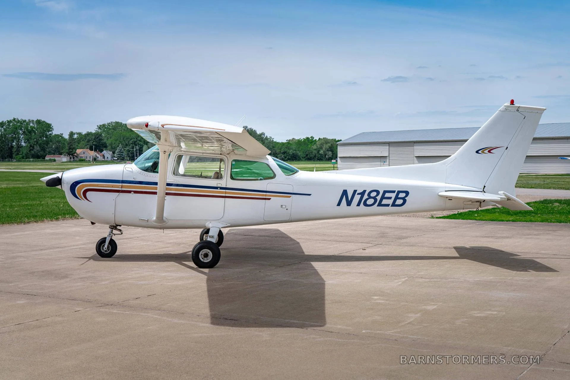 A white Cessna 172 (N18EB) with blue and gold stripes is parked on a tarmac, viewed from the right side.