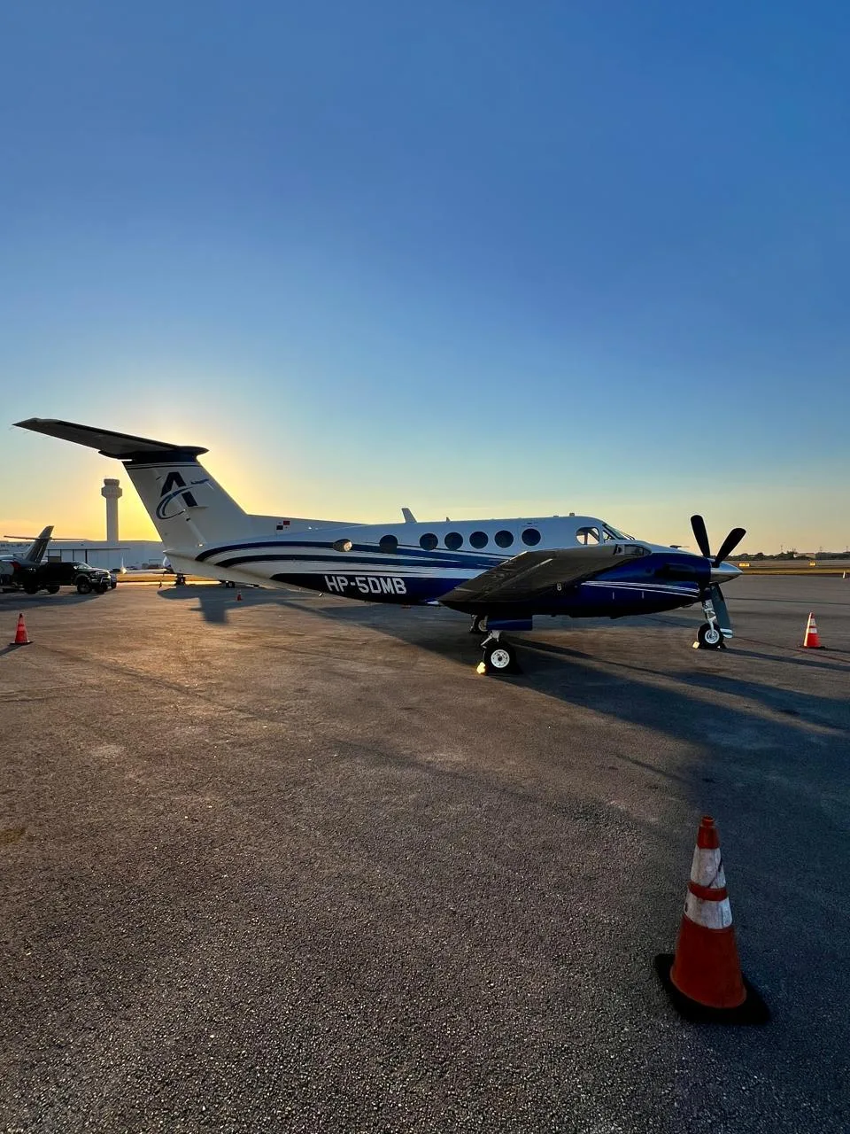 A white and blue turboprop aircraft (King Air) with registration HP-5DMB parked on an airport tarmac at sunset, with an air traffic control tower in the background.