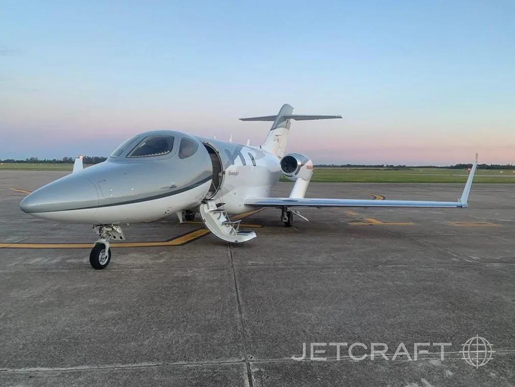 A silver and white HondaJet is parked on an airfield with its main door open at dusk.