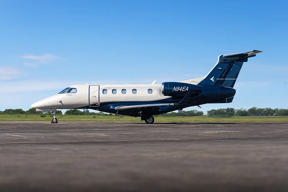 A white and dark blue Embraer Phenom 300E business jet with tail number N84EA parked on an asphalt runway under a clear blue sky.