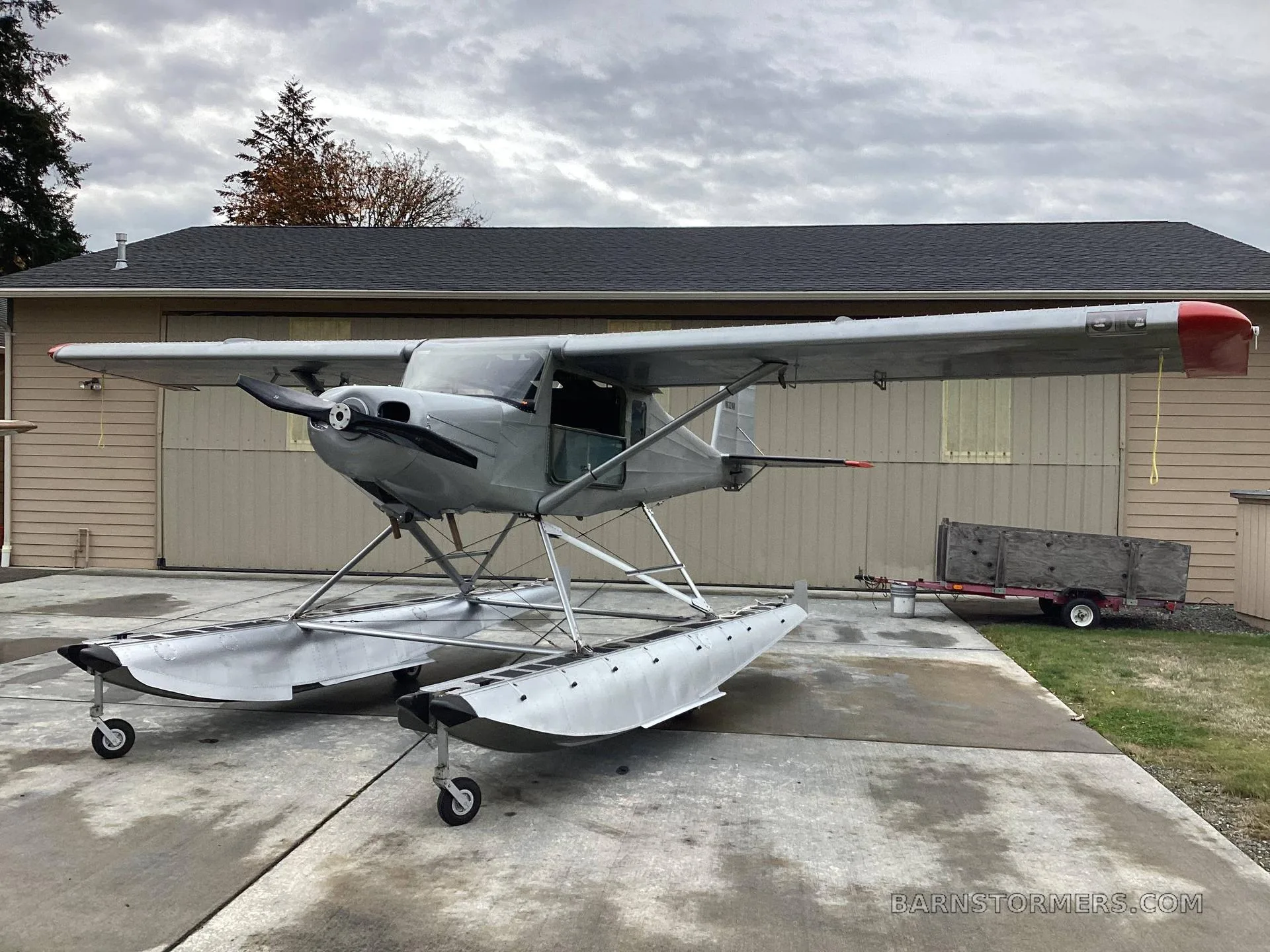 A silver floatplane with a red nose and tail, registration N6324H, parked on a concrete pad in front of a light-colored hangar under a cloudy sky.