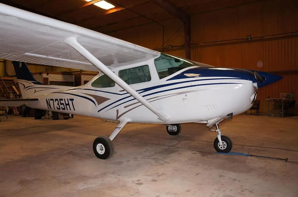A white and blue Cessna 182 Skylane N735HT is parked in a hangar, viewed from the front-right.