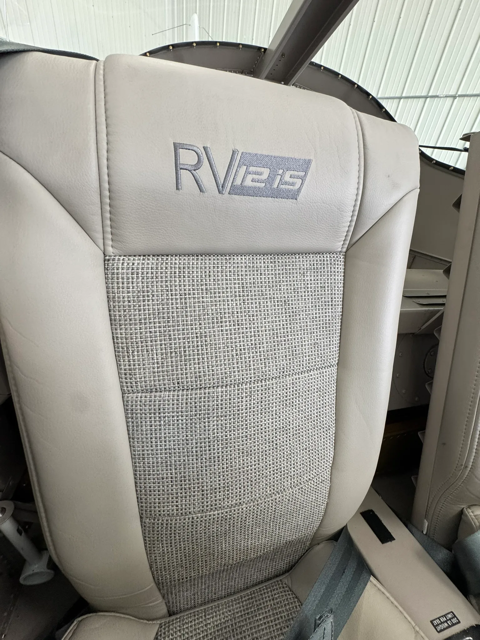 A close-up of an aircraft seat, showing "RV-12iS" embroidered on the beige leather headrest.
