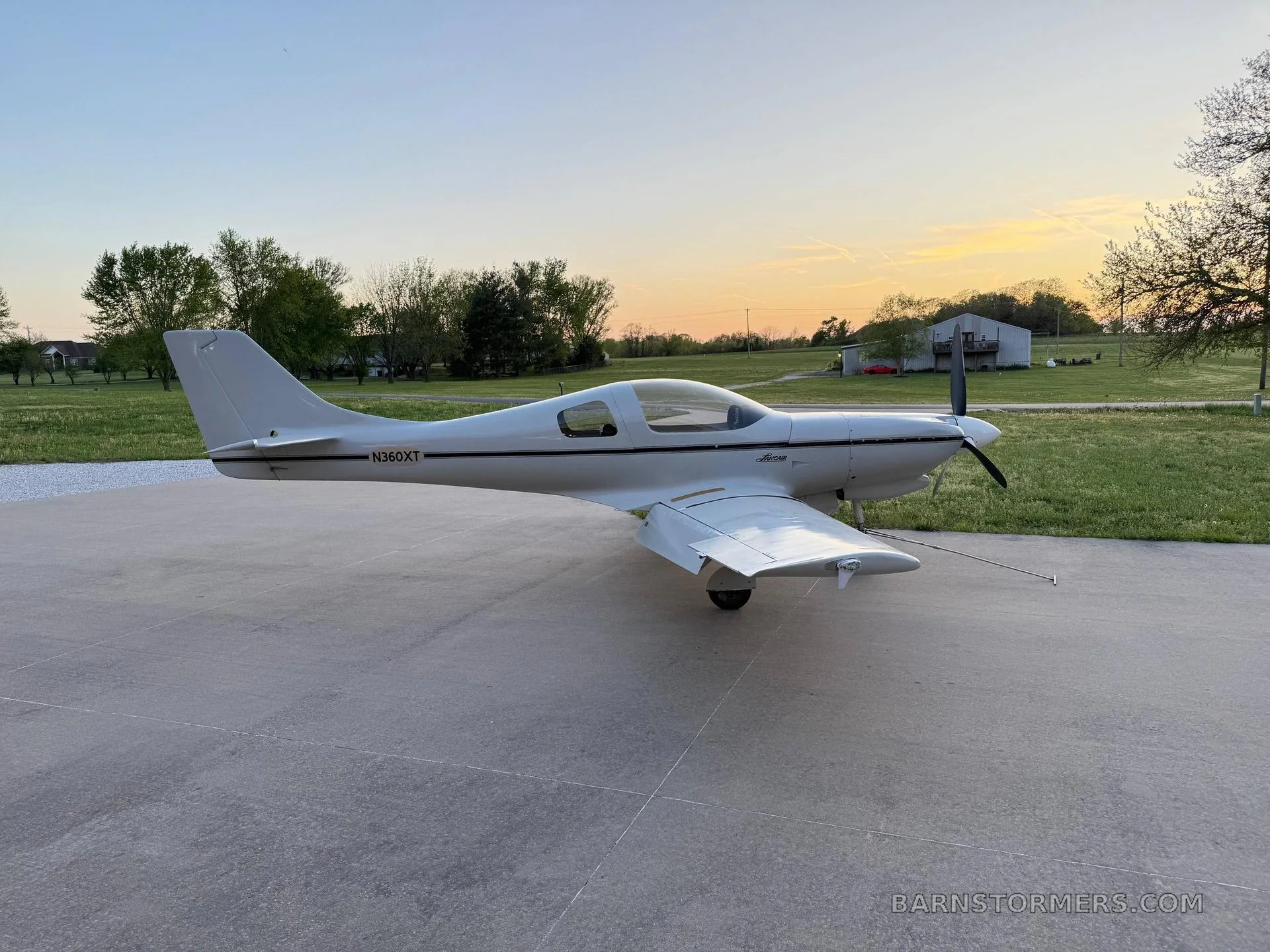 A white Lancair Legacy aircraft (N360XT) with a black stripe is parked on a concrete pad at sunset, viewed from the side.