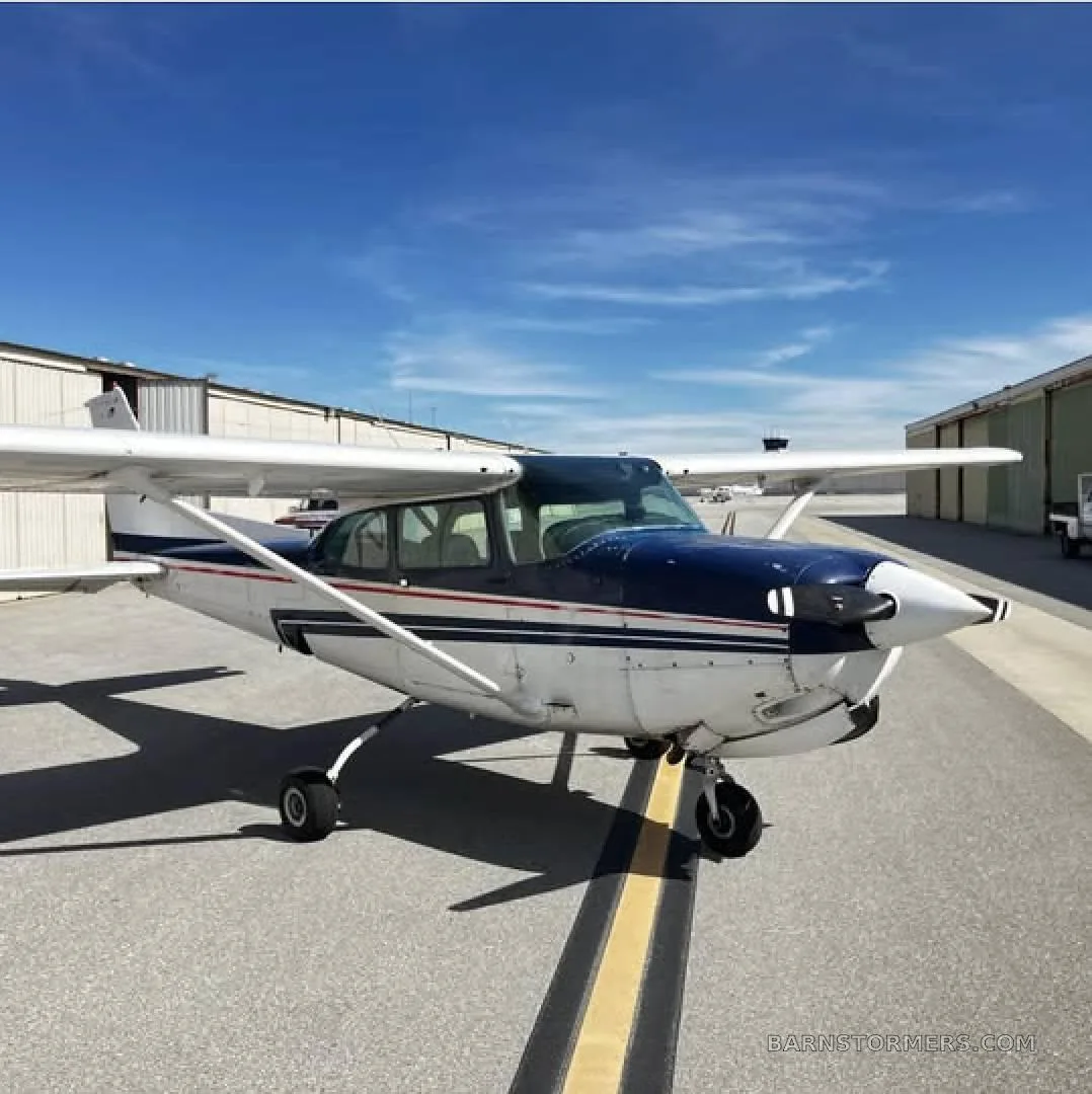 A front-right view of a white and blue single-engine propeller aircraft on an airport tarmac under a blue sky.