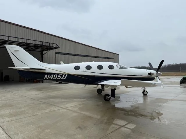 A white and dark blue Daher TBM aircraft, N495JJ, is parked on a wet tarmac in front of a hangar under an overcast sky.