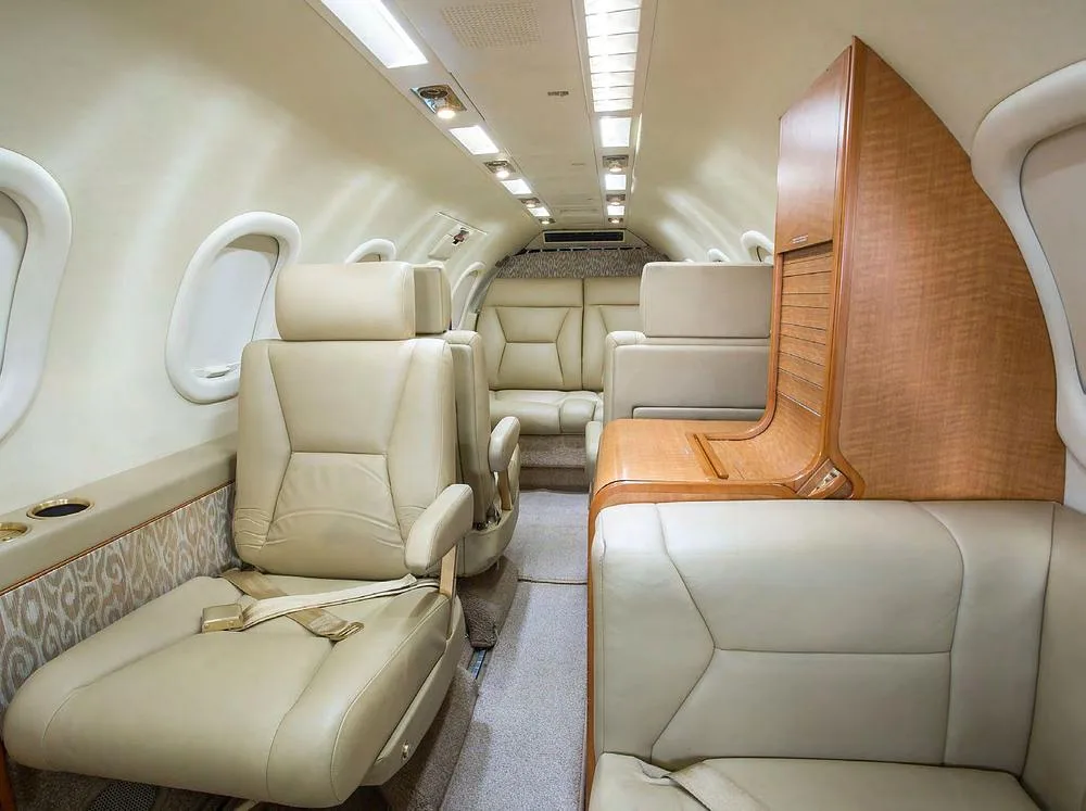 The aircraft cabin interior features light beige leather seats and light wood cabinetry.