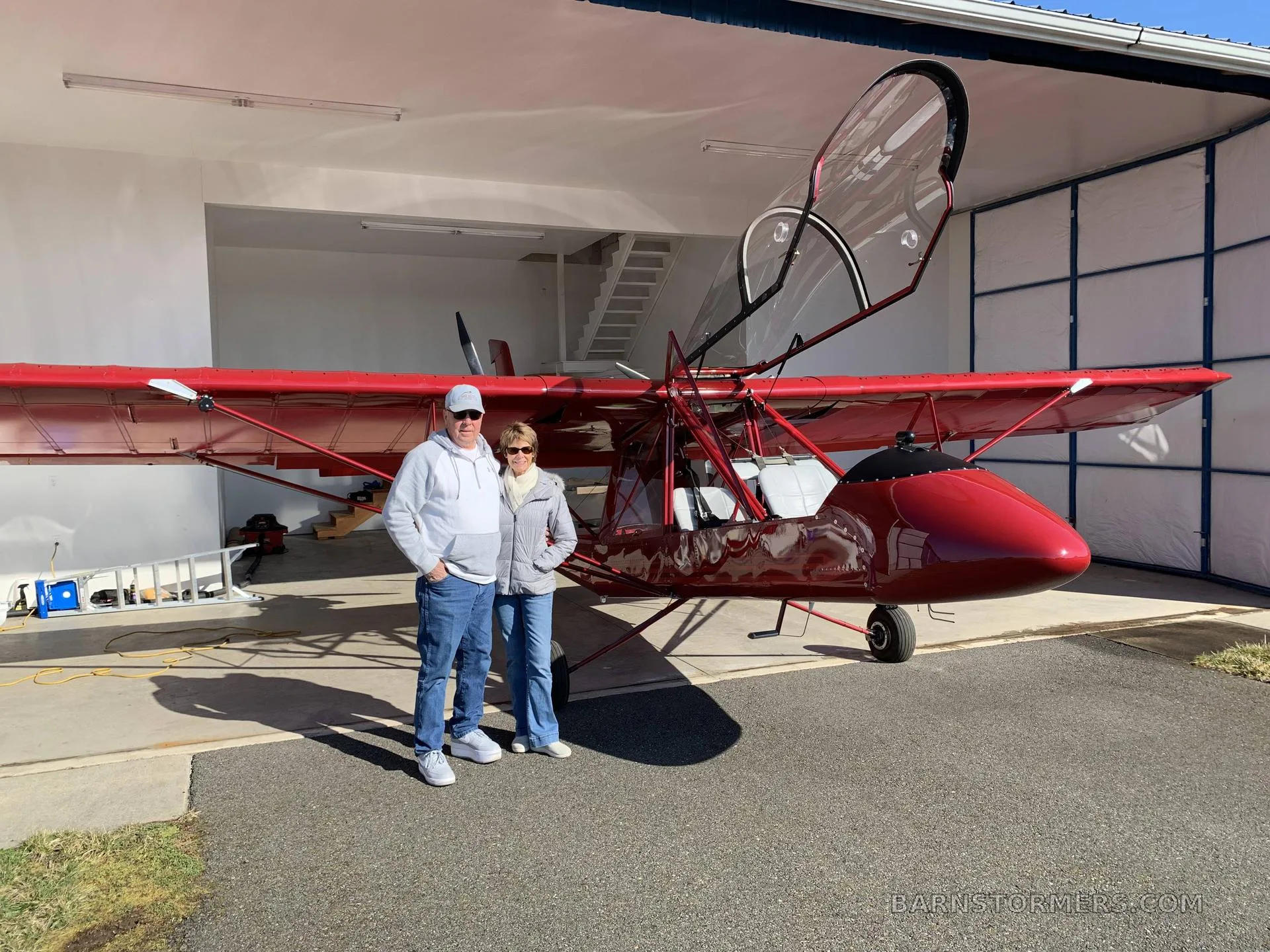 A couple stands smiling in front of a red Air Cam aircraft with its canopy open, parked inside a hangar.