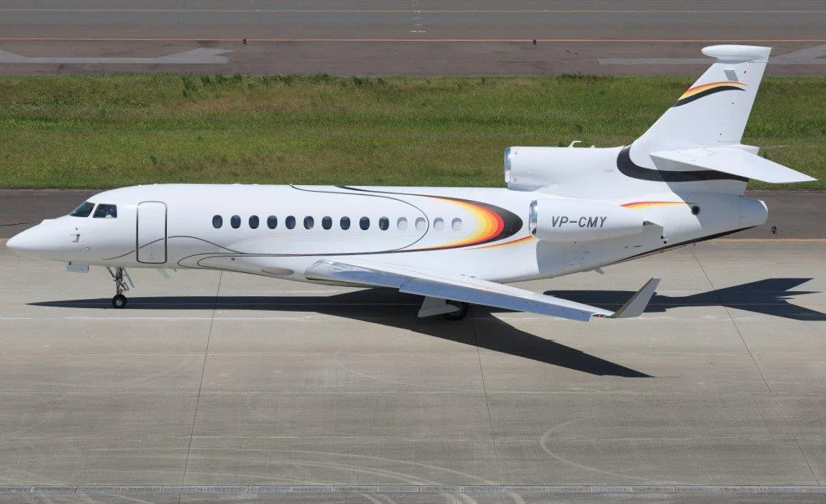 A white Dassault Falcon 900 business jet with a black, red, orange, and yellow stripe livery, registered VP-CMY, parked on an airport tarmac.