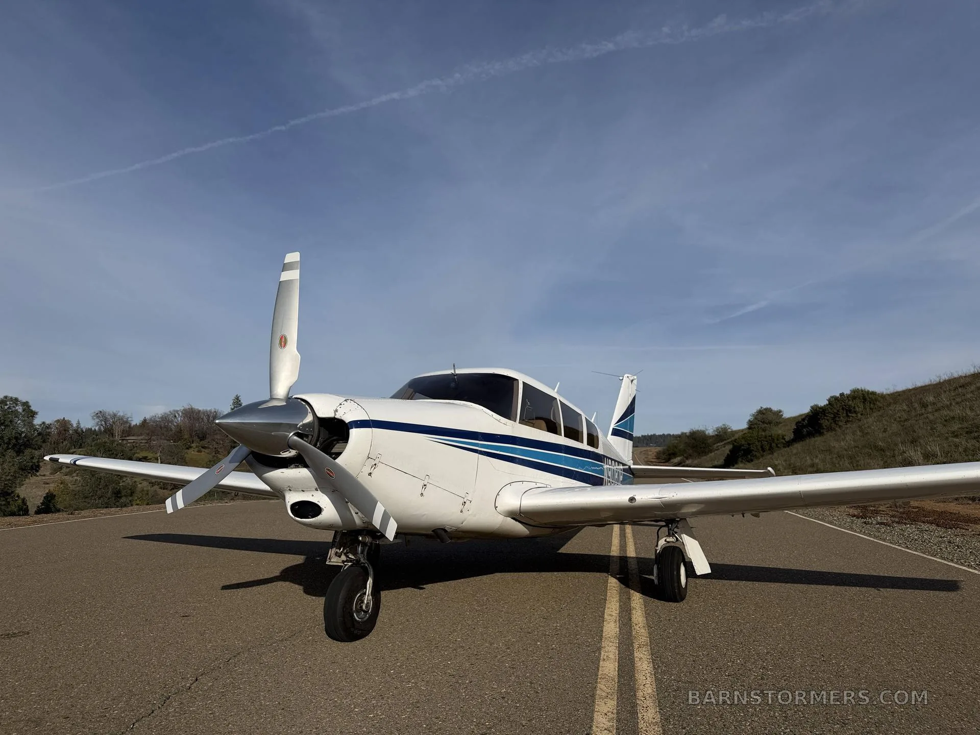 A white single-engine propeller aircraft with blue stripes (N9081P) is parked head-on on an asphalt surface with hills in the background.