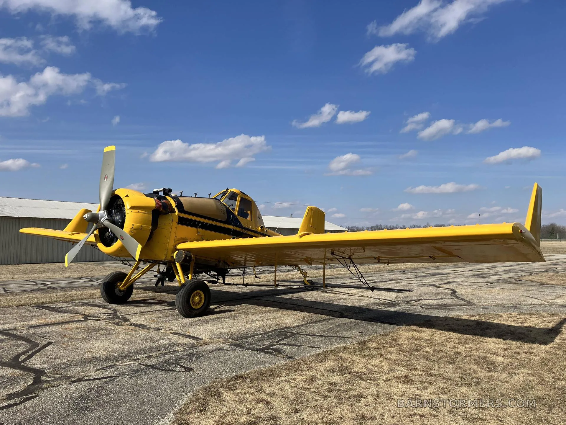 A yellow agricultural aircraft with a black stripe is parked on an asphalt ramp under a blue sky with clouds.