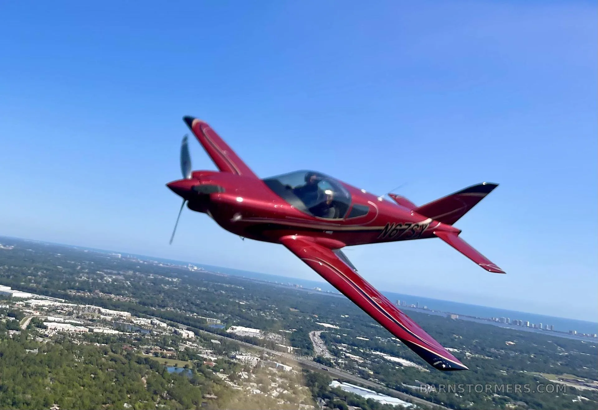 A red single-engine aircraft with registration N67SX flies over a green landscape with buildings and a body of water in the distance.