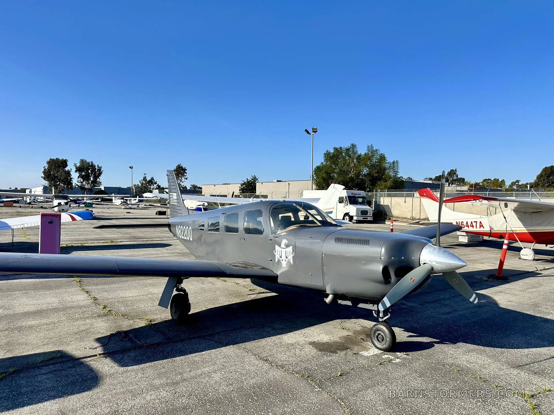 A gray single-engine propeller aircraft, N8220Q, with a white emblem on its nose, parked on a concrete tarmac under a clear blue sky.