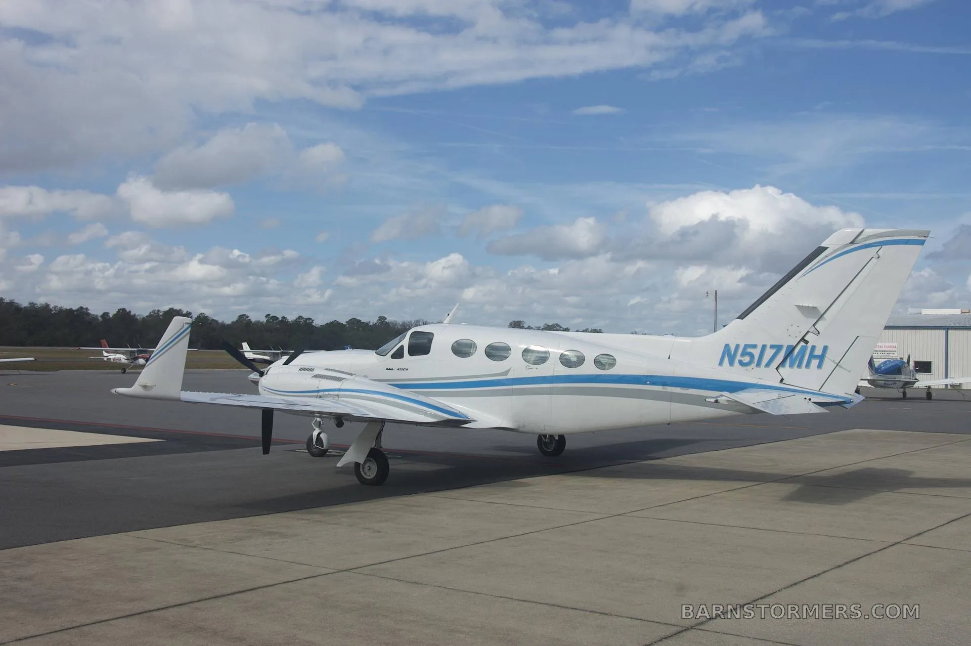 A side view of a white twin-engine propeller aircraft with blue and grey stripes (N517MH) parked on a tarmac with other aircraft in the distance.