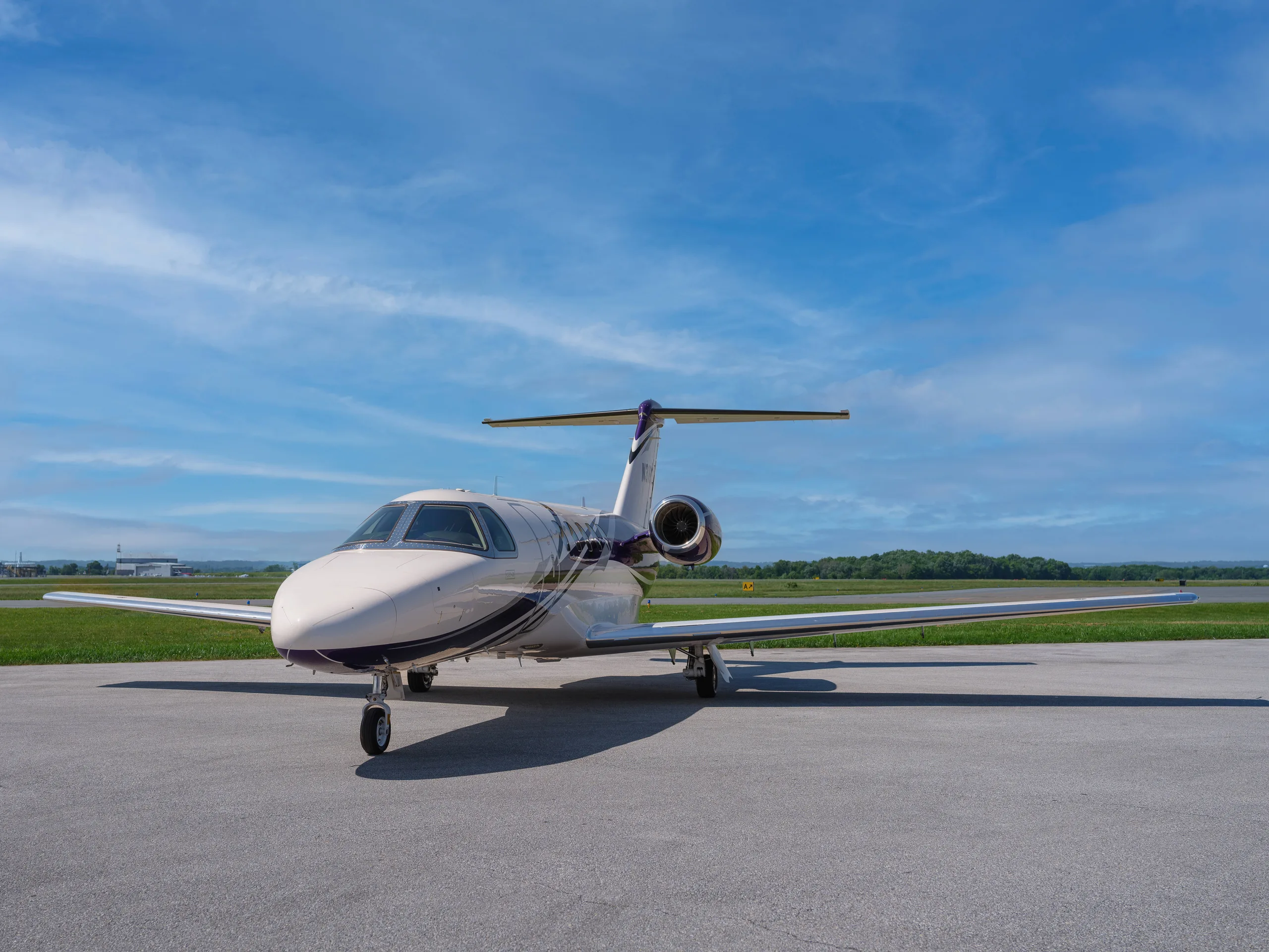 A white and purple Cessna Citation CJ4 business jet is parked on an asphalt tarmac under a blue sky with scattered clouds.