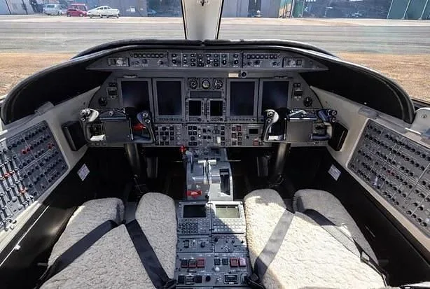 The cockpit of a private jet features a complex instrument panel, multiple screens, and two pilot seats with sheepskin covers.