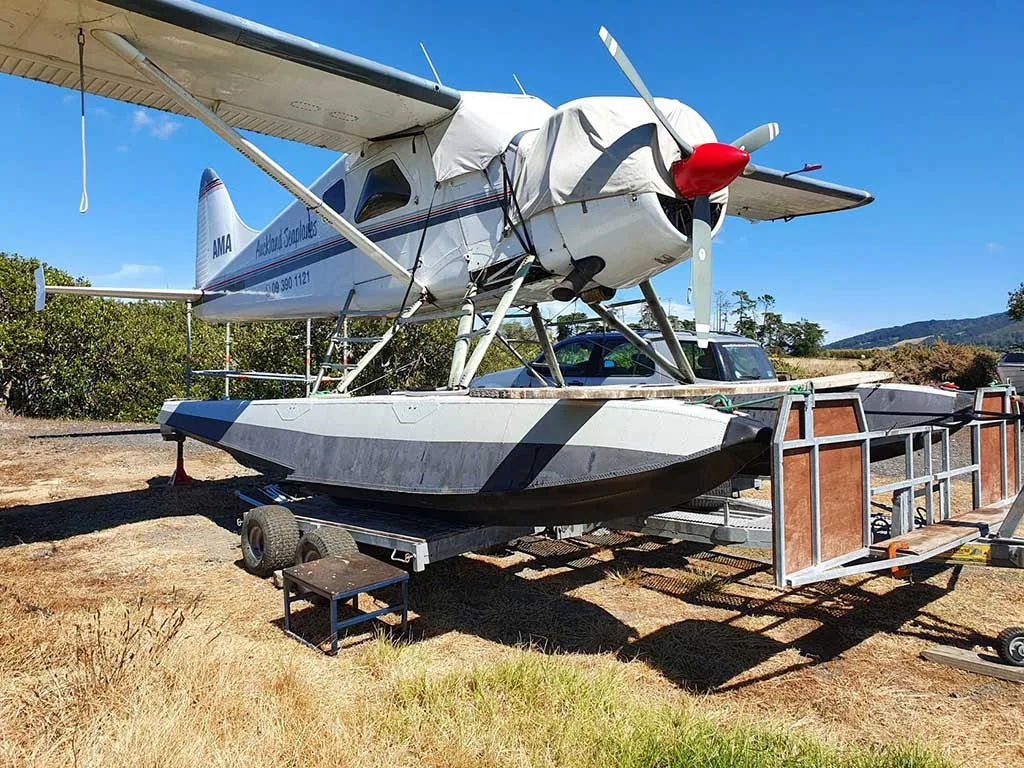 A white seaplane with a red propeller spinner and grey floats is parked on a trailer in a grassy area under a clear blue sky.