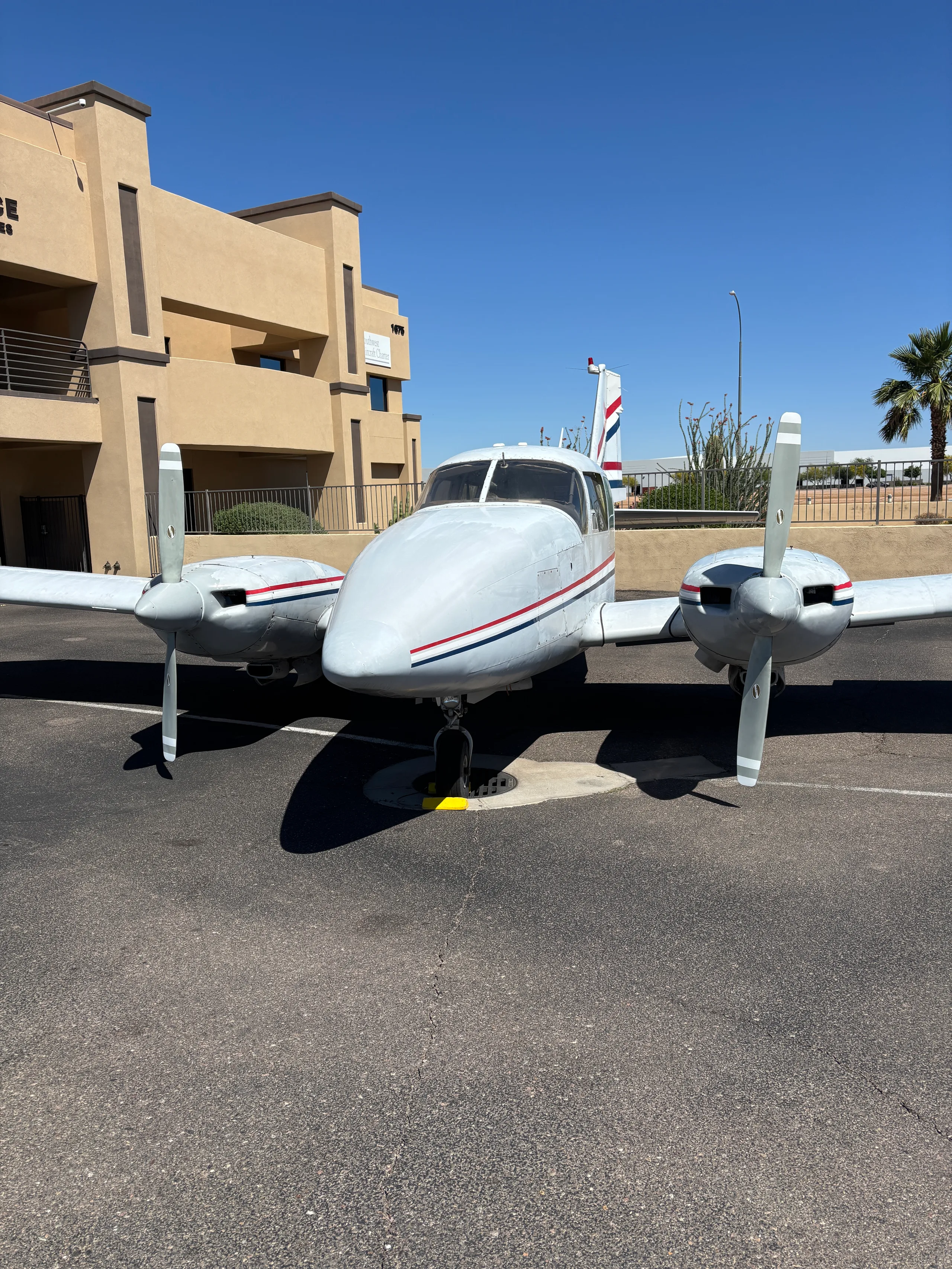 Front view of a light gray twin-engine propeller aircraft with red and blue stripes, parked on an asphalt lot.